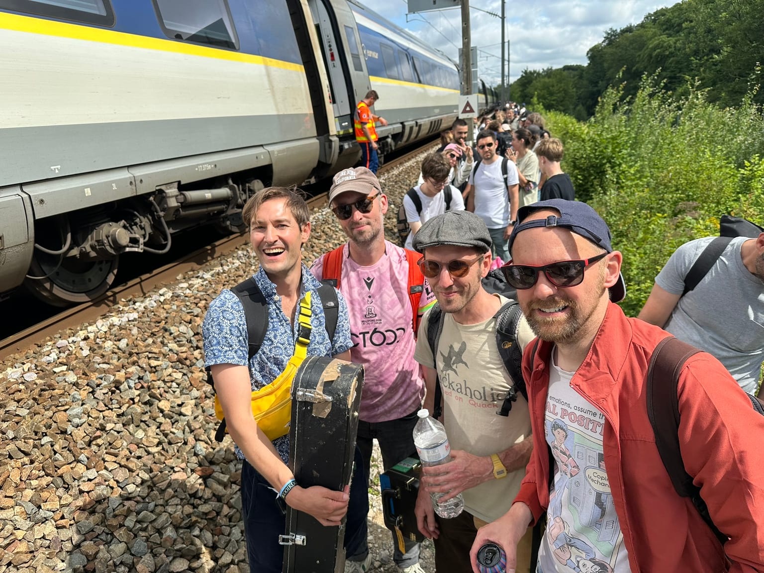 Four men carrying instrument cases stand on the ballast beside a stopped Eurostar train, a long line of other passengers walking along the track behind them