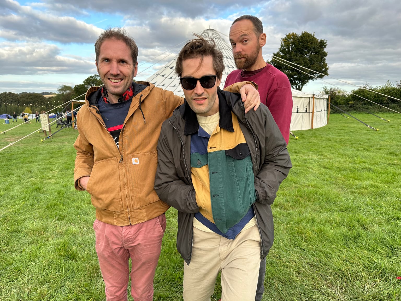 Three men standing arm in arm in a festival field, a canvas tent and strings of lights behind them