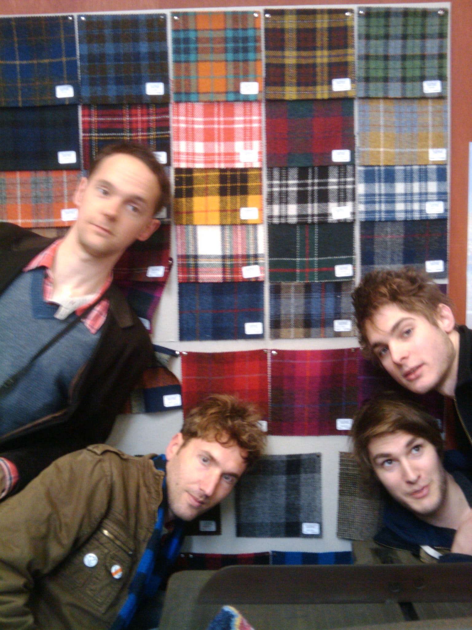 Four members of the band leaning into the camera in front of a wall hung with dozens of tartan and tweed cloth samples