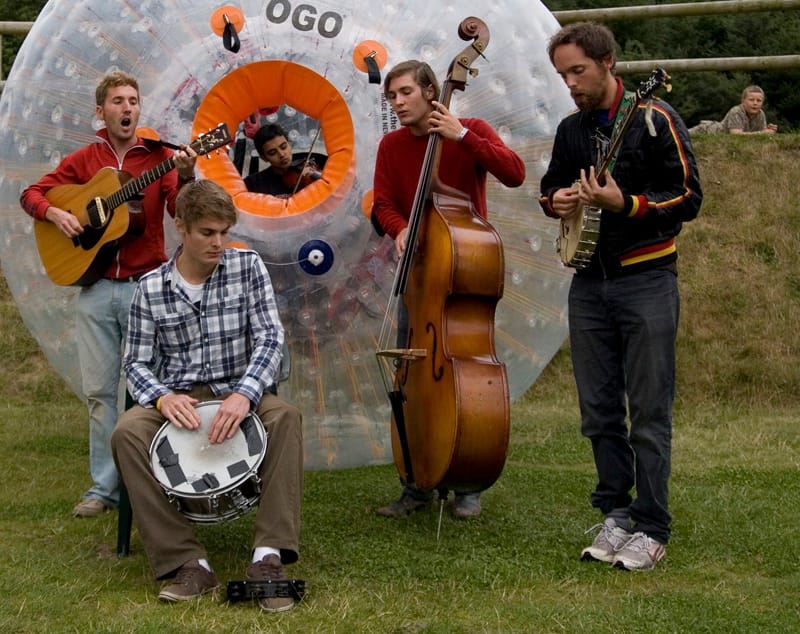 Five musicians playing guitar, snare drum, double bass and banjo on grass in front of a large inflatable zorb ball, with a fifth member playing violin inside it